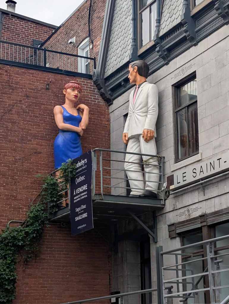 Die Skupturen einer Frau und eines Mannes stehen auf einem Balkon und unterhalten sich.