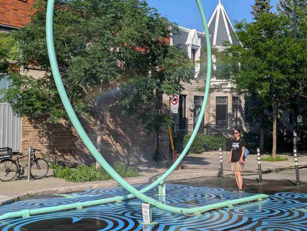 Ein Spray-Pool in Form einer Schleife, der Wasser versprüht; ein Regenbogen hat sich dazwischen gebildet. Eine Frau steht rechts daneben und lacht.