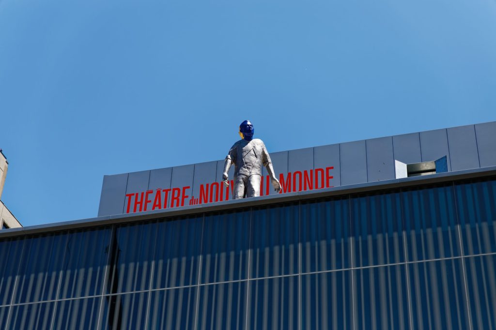 Auf einem Theaterdach steht eine silberne männlich aussehende Figur.