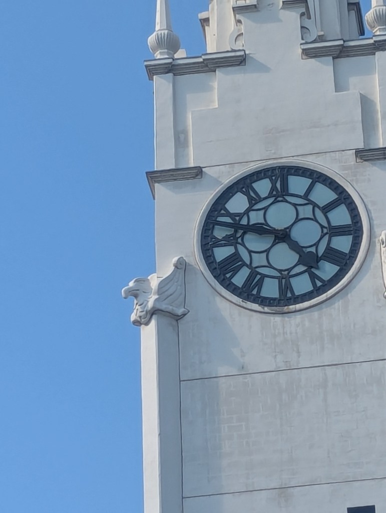 Eine weißer Turm mit einer Uhr darauf. Links am Turm ist eine Plastik in Form eines Adlerkopfes angebracht.